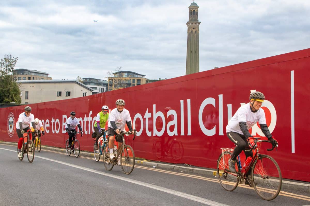 A row of Cyclists cycling in front of a red hoarding with 'Welcome to Brentfor ootball Club' printed on it