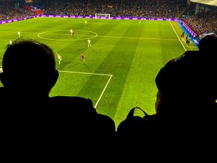Silhouette of two football fans watching a floodlit game from high in the stands